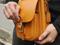 職人手作皮革皮扣直式單肩包，呈現溫潤的芥末黃色。包身由厚實的植鞣皮革製成，縫線精緻，質感紮實。包蓋上設有金屬插扣，方便開闔，側面有兩個小口袋，整體風格簡約而復古。