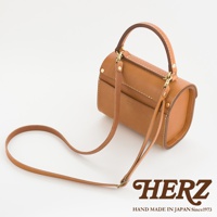 職人手作皮革復古杜勒斯手提包，採用棕色牛皮製成，帶有圓潤的桶狀造型、頂部提把和可拆卸斜背帶。包身設有雙層前袋，以白色車線縫合，並有金屬釦件裝飾。