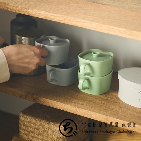 一隻手正從木質層架上拿取一個淺藍色的美濃燒茶壺，旁邊疊放著兩個薄荷綠色的茶壺，右側有一個圓形蓋盒，層架下方印有宇治田原製茶場直賣部的品牌標誌與文字。
