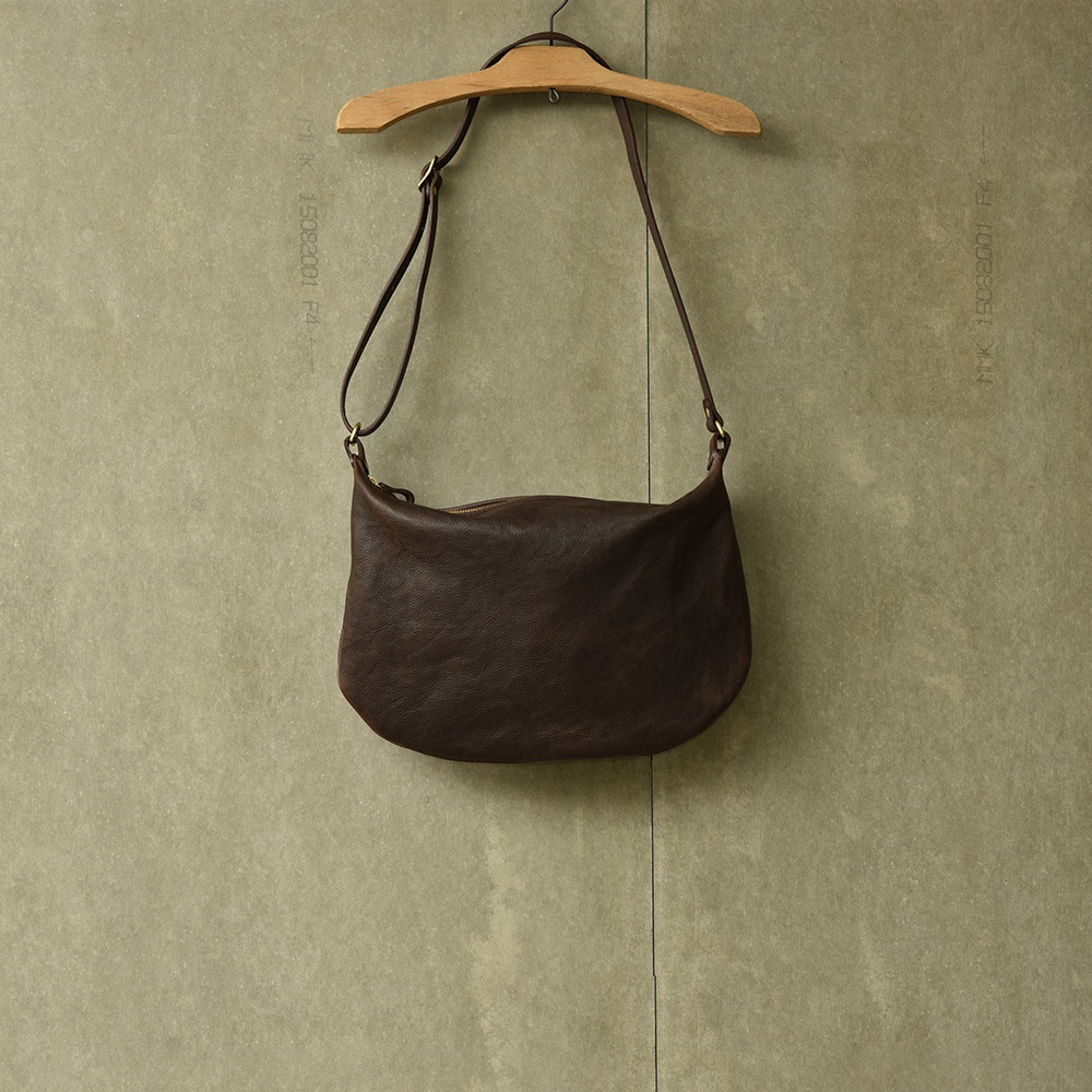 職人手作皮革Organ單肩半月包，深棕色，圓潤彎月造型，頂部有拉鍊開口，配備可調節長度的皮革肩帶，透過木製衣架懸掛在淺綠色背景前。