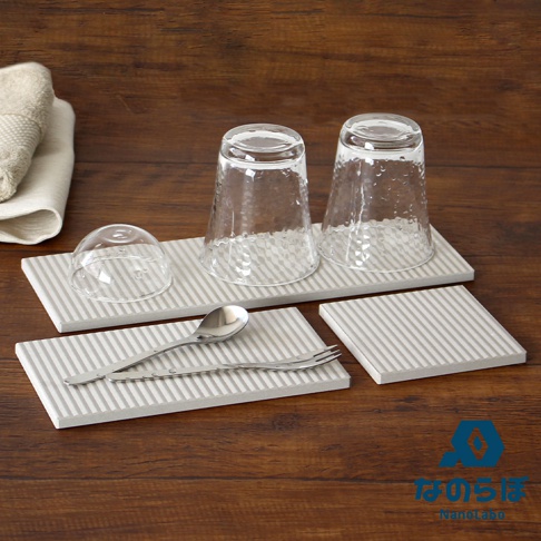 淺灰色條紋珪藻土置物墊，上面擺放著兩個透明玻璃杯、一個玻璃蓋、一把湯匙和一把叉子，背景為木質桌面，左側有一塊摺疊的毛巾。