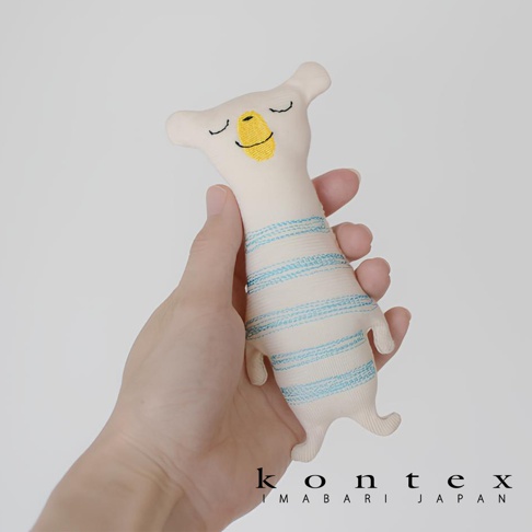 手持一個米白色有機棉材質的可愛動物造型抓握玩偶，玩偶有著閉眼微笑的表情，身上繡有藍色條紋，底部有四隻小腳。