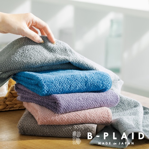 一疊五色（深灰、粉紅、淺紫、淺藍、淺灰）的今治毛巾，表面有鱗紋織紋，由手拿起最上方的淺灰毛巾。