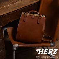 棕色皮革復古口金後背包，具備頂部提把、可調節式肩帶、金屬扣環及四個角落的加固設計，擺放在椅子上，旁邊是木製桌子。