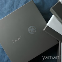 深灰色紙盒，印有銀色「Tsudoi」字樣及花形圖案，旁邊疊放著兩個黑白相間的紙盒。