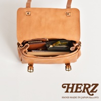 一個淺棕色皮革製作的復古郵差包，包包內裝有多個皮夾和卡夾，包包上方蓋子敞開，露出內部細節，包包正面有兩個金屬扣環。