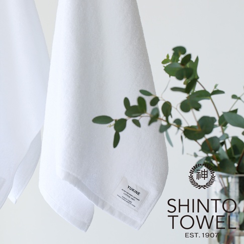YUKINE 雪中漫步有機棉蓬厚迷你浴巾，白色，材質為 100% 棉。浴巾蓬鬆柔軟，邊緣縫有品牌標籤，一旁擺放著綠葉裝飾，整體呈現簡約純淨的風格。