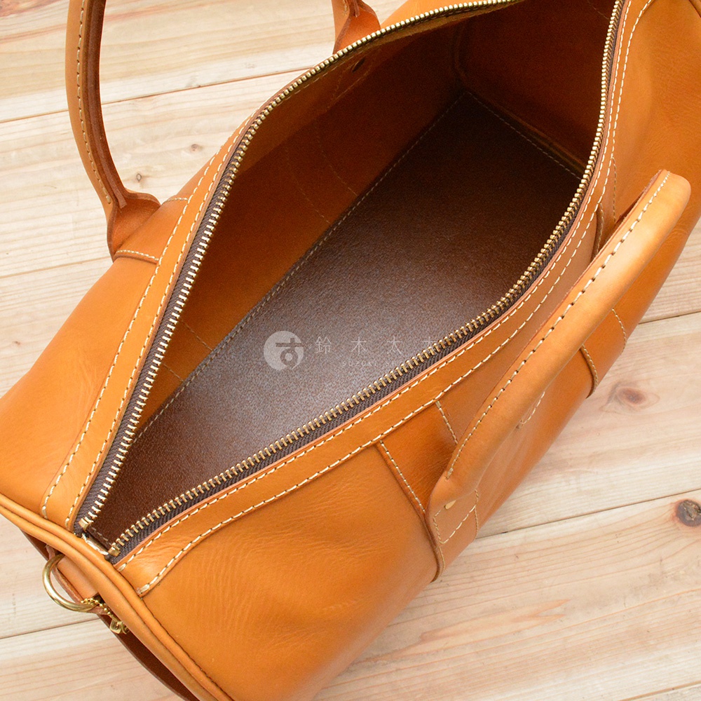 職人手作皮革肩背手提拉鍊波士頓包，採用天然皮革材質，呈現溫潤的淺棕色調。包款設計簡潔，具有拉鍊開口與提把，內部空間寬敞，可容納多樣物品。車線細緻，展現職人手工的質感。