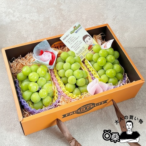 橙色禮盒內盛有三串飽滿翠綠的日本麝香葡萄，葡萄果實圓潤，色澤鮮亮，擺放在刨絲紙和彩色網格襯墊上。禮盒一側裝飾著粉色和紅色的仿真花朵，並附有一張印有日文和卡通人物的卡片。