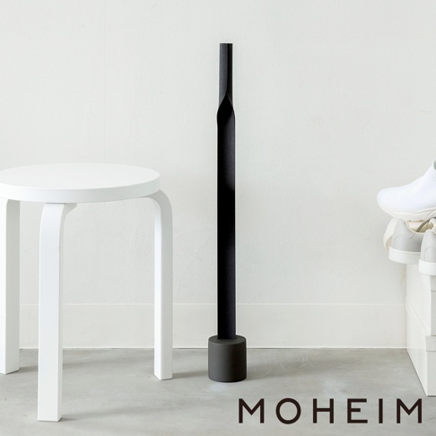 黑色金屬鞋拔，配有圓形底座，立於白色牆壁旁的灰色地板上，旁邊有一個白色凳子和一雙白色運動鞋。