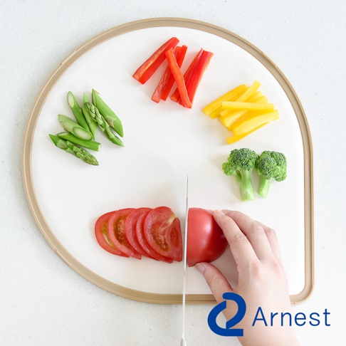 淚滴型白色抗菌料理砧板，邊緣有淺棕色框架。砧板上擺放著切塊的紅甜椒、黃甜椒、蘆筍、花椰菜和切片的番茄，其中一個番茄正被刀切開。