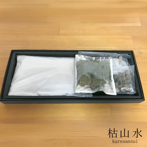 黑色禮盒內裝有日本枯山水模型組件，包含一包白色細沙、一包乾燥青苔、數顆灰色碎石，以及一支木製迷你耙子，畫面右下方標註有枯山水及 karesansui 字樣。