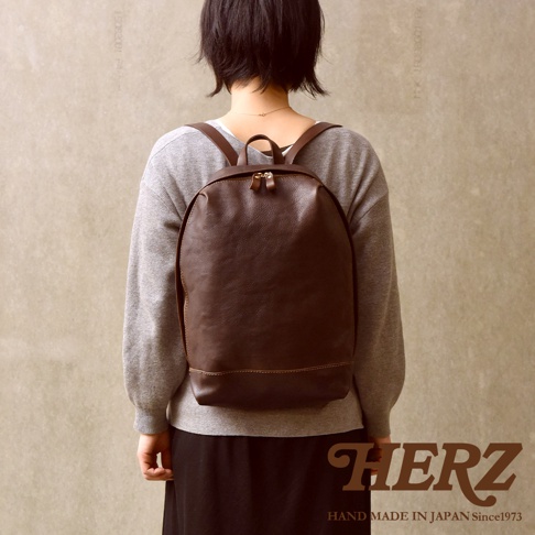 一位留著短髮的女性背著日本HERZ品牌的手工職人後背包，該包款由深棕色質感皮革製成，呈現簡約俐落的圓弧褶皺造型，並附有金色拉鍊，整體風格知性優雅，右下角印有HERZ及日本手工製造文字。