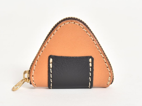 職人手作皮革零錢包，呈三角飯糰造型，主體為淺棕色皮革，中央區域為黑色皮革拼接，邊緣以白色縫線裝飾，並配有金色拉鍊。