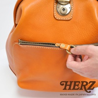 職人手作皮革輕旅行後背包，橘色皮革材質，帶有黃銅色拉鍊和圓形拉環，特寫拉鍊細節，底部有品牌標誌HERZ。