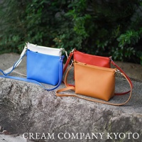 三個京都職人手作隨身手機包，分別為亮藍色、紅色與橘棕色，皆為簡約方正的設計，配有可調式細長肩帶與金屬扣環，展現皮革質感。