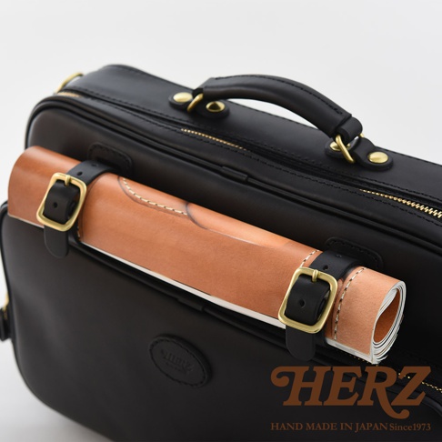 黑色皮革兩用商務公事包，附有可固定捲狀物品的黑色皮革束帶及黃銅扣環。包款頂部有手把，側邊有拉鍊。