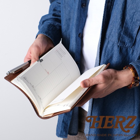職人手作皮革B6記事本書套，展示棕色皮革材質，內頁為橫線格式的行事曆，由雙手捧持，左手持筆。