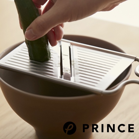 銀色不鏽鋼雙刃切片器，有兩條平行切槽，置於棕色碗上，一隻手正將小黃瓜放入切槽中。