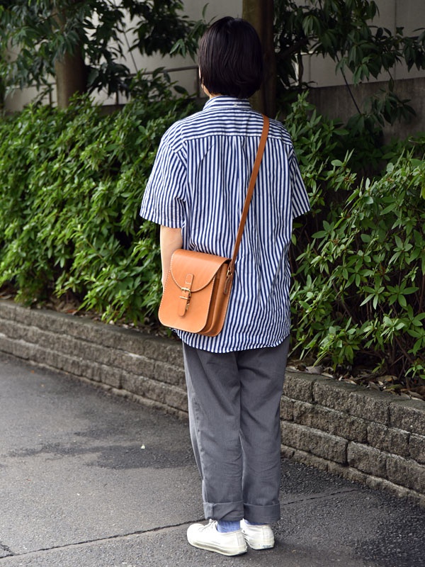 棕色皮革翻蓋迷你單肩包，配有金色搭扣和可調式肩帶，斜挎於穿著藍白條紋襯衫和灰色長褲的人身上。