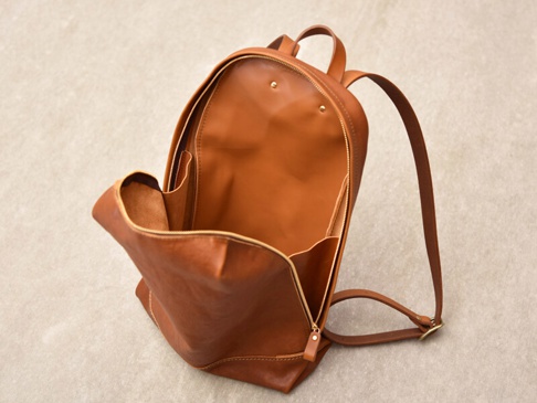 職人手作皮革後背包，採用簡約褶皺設計，顏色為溫暖的棕褐色。材質為真皮，質感細緻，包身敞開，露出內部隔層，肩帶為可調式設計。