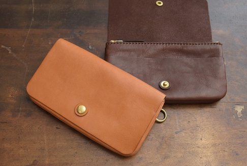 淺棕色皮革長夾，附有黃銅按扣和掛環；深棕色皮革長夾，上方敞開，露出拉鍊開口和內層，同樣配有黃銅按扣。兩者皆為手作皮革製品。