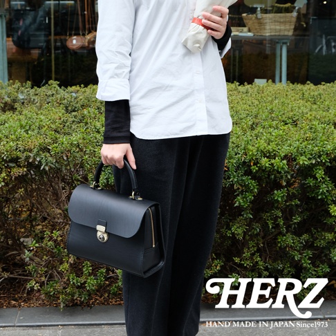 日本HERZ手工皮革手提包，附有金色鎖扣和拉鍊，由黑色皮革製成，搭配白色襯衫和黑色長褲，背景是綠色植物。