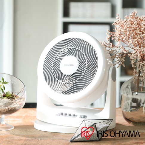 白色IRIS OHYAMA PCF-HM23 空氣循環扇，圓形扇網上有螺旋紋路，底部有按鈕，擺放在木質桌面上，背景為書架與乾燥花飾。