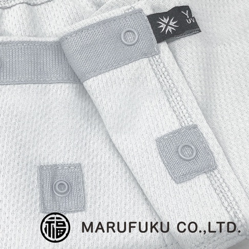 這是一張近距離特寫照片，呈現 MARUFUKU 丸福纖維 YAKENU 遮陽面罩的細節，材質為淺灰色且具有細緻紋理的機能布料，上方縫有一枚帶有太陽標誌的品牌標籤，邊緣處設有三個塑膠按扣，用於調整配戴方式，左下角則印有 MARUFUKU CO.,LTD. 的品牌名稱及其特有的福字圓形標誌。