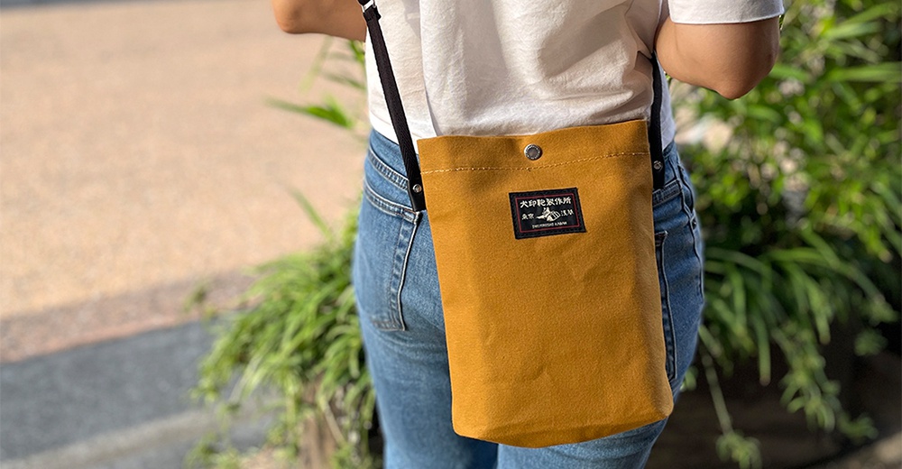 一位女士身穿白色T恤和牛仔褲，斜挎一個薑黃色帆布側背包，包上有一個黑色標籤，上面印有日文文字和圖案。