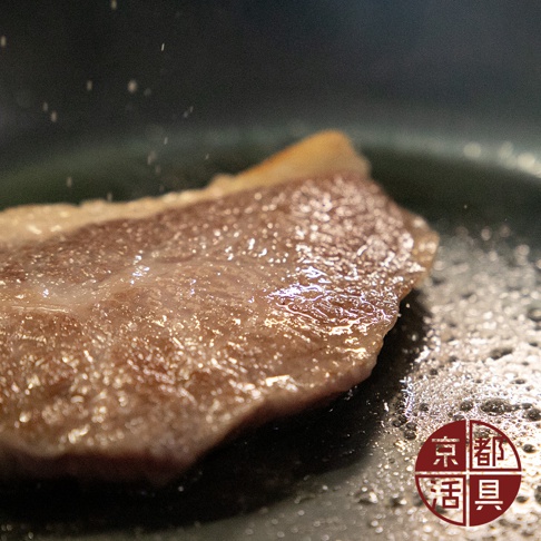 一塊褐色的肉正在烹煮，表面有油光，灑上了白色顆粒。一旁是冒著細小泡沫的鍋底，角落有紅色圓形標誌。
