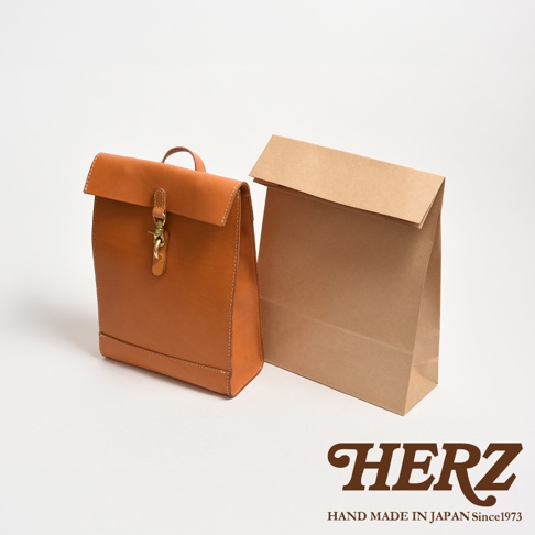 棕色皮革後背包，設計靈感來自紙袋，搭配金屬扣環，旁邊另有一個紙袋。
