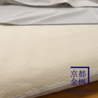 淡黃色絎縫紋理的單人毯，上方疊著一層淺灰色毯子，右下方有藍色邊框的金色日文字樣和英文品牌標誌。