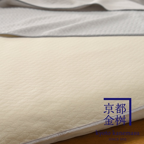 淡黃色絎縫紋理的單人毯，上方疊著一層淺灰色毯子，右下方有藍色邊框的金色日文字樣和英文品牌標誌。