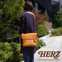 日本HERZ皮革雙扣旅行郵差包，經典雙插釦設計，駝色皮革展現職人手作質感，肩背於身後，背景為綠意盎然的植物與現代建築。