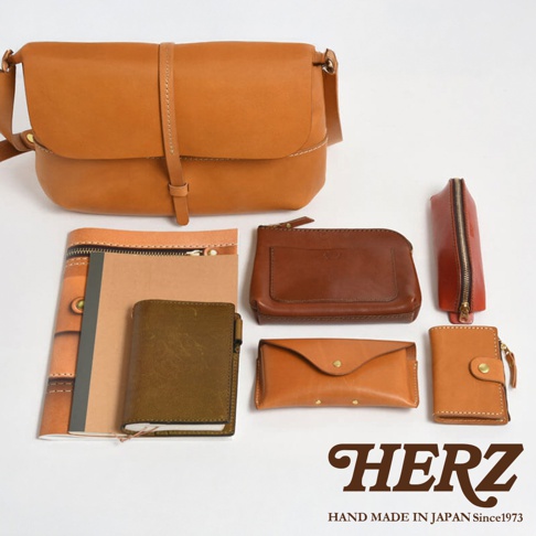 日本HERZ品牌手工製作的皮革產品，包含一個棕色斜背包、幾個小皮件如零錢包、卡夾，以及一本筆記本，背景為白色。