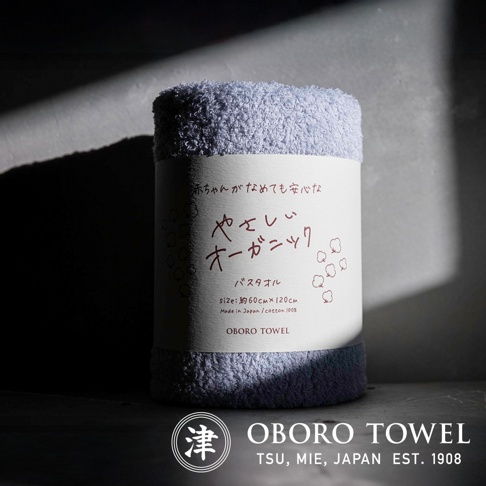 一條淺藍色的有機棉浴巾，質地柔軟，捲起後標籤上印有日文和英文說明「やさしいオーガニック バス タオル」、「SIZE: 約60CM×120CM」、「Made in Japan / cotton 100%」及「OBORO TOWEL」字樣。