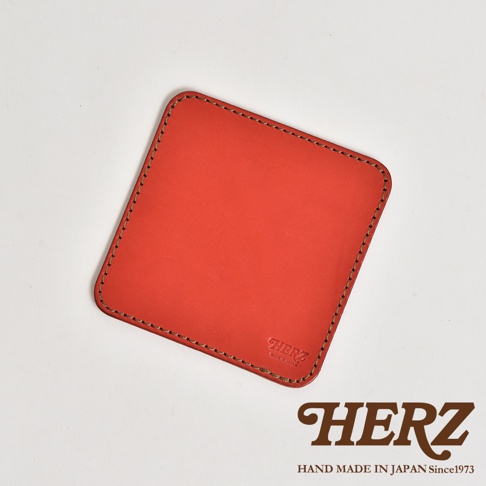 這是一款由日本品牌HERZ職人手工製作的紅色方形皮革墊，選用硬挺的Latigo皮革打造，邊緣以細緻的縫線收邊。墊片右下角壓印有HERZ品牌標誌，下方並註明MADE IN JAPAN字樣。整體設計簡約厚實，呈現優雅的職人工藝質感。