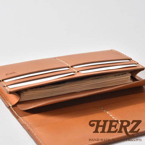 這是一款日本HERZ手工製作的皮革長夾，呈現淺棕色調。畫面以近距離俯視角度展示長夾打開後的內部結構，清晰可見多個卡槽層次以及存放於主夾層內的紙鈔，皮革質感細膩且縫線工整，右下角印有HERZ品牌標誌與手工製造資訊。