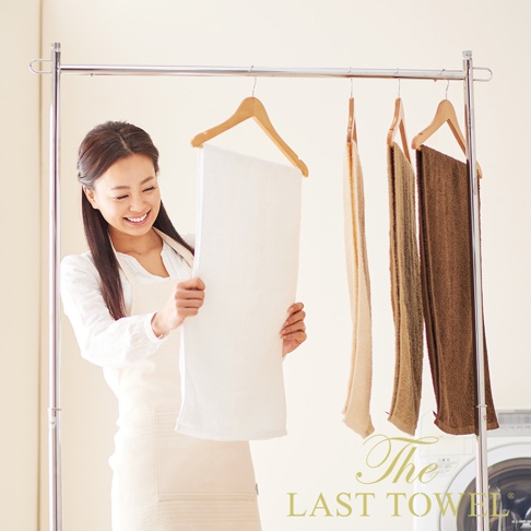 一位女士展示一條白色純棉柔毛巾，旁邊掛著另外三條不同深淺的棕色毛巾，所有毛巾皆在衣架上晾曬。