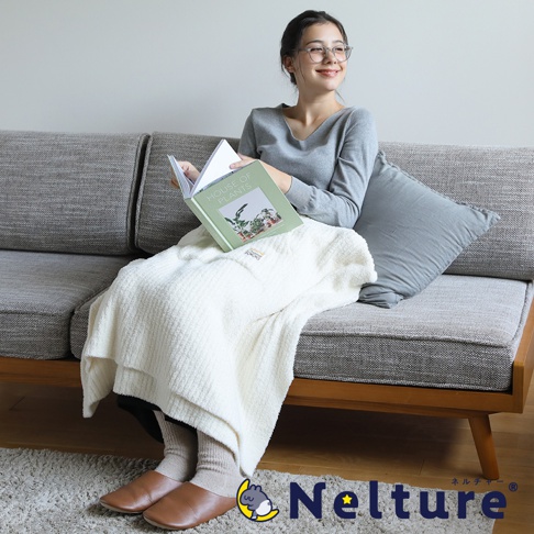 一位女士在沙發上閱讀，身上蓋著一件奶油色的斗篷，腳上穿著皮革拖鞋。