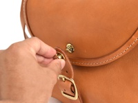 手正在操作一個淺棕色皮革馬鞍包的黃銅旋轉鎖扣，包包有明顯的縫線細節。