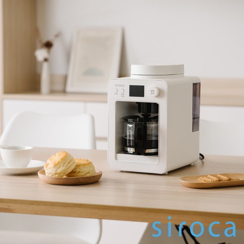 白色 SC-A3520 自動研磨咖啡機，配有玻璃咖啡壺、黑色研磨器和一個小型顯示螢幕。咖啡機放置在木質桌面上，旁邊有兩塊司康餅和一杯咖啡。