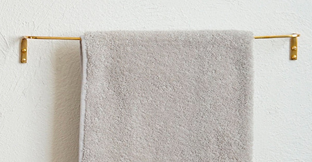 輕奢黃銅單桿毛巾架，黃銅材質，簡潔的單桿設計，固定於白色紋理牆面上，懸掛一條淺灰色毛巾。