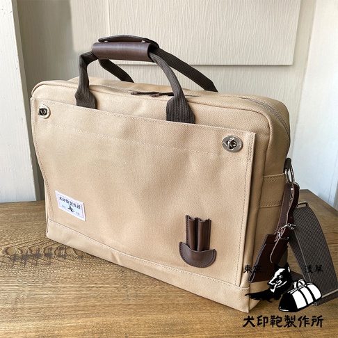 犬印純綿帆布商務公事包，米色帆布材質，咖啡色皮革提把與裝飾，B4尺寸，配有可拆卸肩帶，正面有品牌標籤與皮革筆插設計，採用金屬旋轉釦開合。