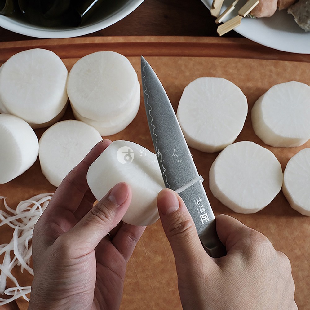 一把銀灰色金屬主廚刀，刀刃有波浪紋，正在切片白蘿蔔。周圍散落著已切好的白蘿蔔圓片，以及部分切下的細絲。