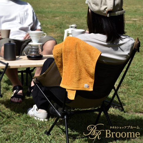 橘黃色今治純棉掛脖毛巾，紋理清晰，掛在露營椅上，旁邊有兩人正在戶外享受咖啡時光。