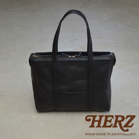 黑色皮革手提包，方形設計，頂部拉鍊開合，設有雙提把，包身正面有一個貼袋，材質為粒面皮革。