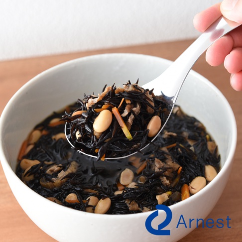 一只手拿著不鏽鋼瀝水湯匙，湯匙中裝滿了黑色的髮菜、白色的豆子和切碎的紅蘿蔔，湯匙上方有湯汁滴落，下方是裝有相同食材的白色碗。