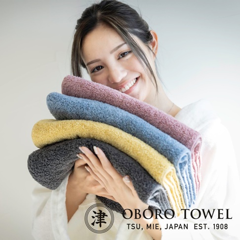 五條不同顏色的毛巾堆疊，包括深灰色、黃色、藍色和粉紫色，質地柔軟蓬鬆。